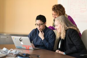 three women beside table looking at MacBook. Christian business leader guiding a team with integrity and servant leadership, reflecting faith-based values in a modern workplace