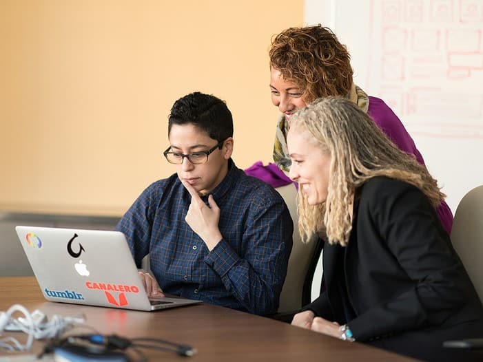 three women beside table looking at MacBook. Christian business leader guiding a team with integrity and servant leadership, reflecting faith-based values in a modern workplace